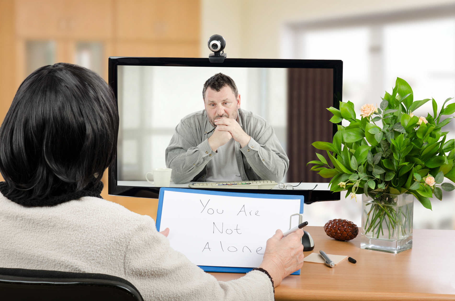 Middle aged man sitting in the monitor talks with psychotherapist via online video chat. He looking depressed. Black-haired psychiatrist holds written message for him - You are not alone. Horizontal shot indoors